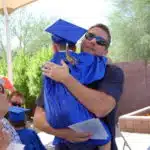 A man in a graduation cap and gown is hugging a young boy at an early learning center. A man in a graduation cap and gown is hugging a young boy at an early learning center.