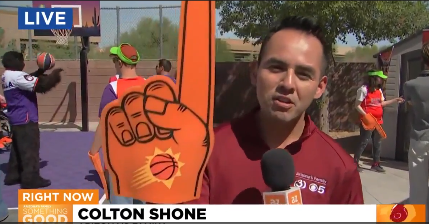 A news reporter holds a microphone and a large foam finger with a basketball logo at an outdoor basketball event, with children playing in the background.