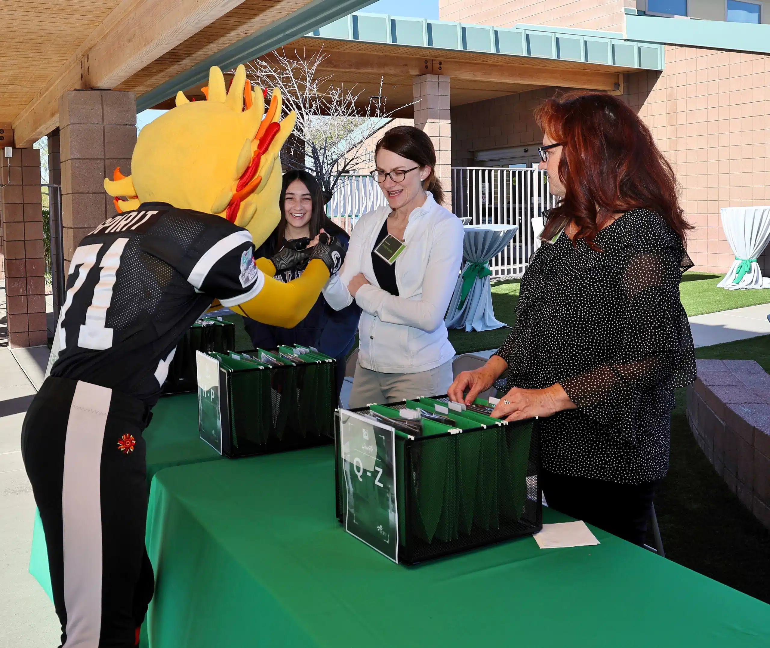 A person in a mascot costume greets two women at an outdoor event registration table, while another woman stands nearby smiling. A person in a mascot costume greets two women at an outdoor event registration table, while another woman stands nearby smiling.