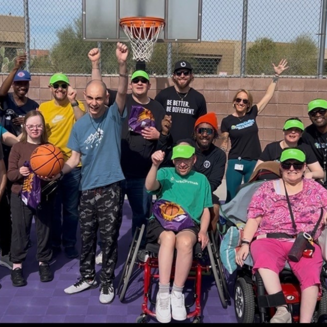 A diverse group of people, some in wheelchairs, pose and smile on an outdoor basketball court, with a hoop visible in the background. Several participants wear green caps.