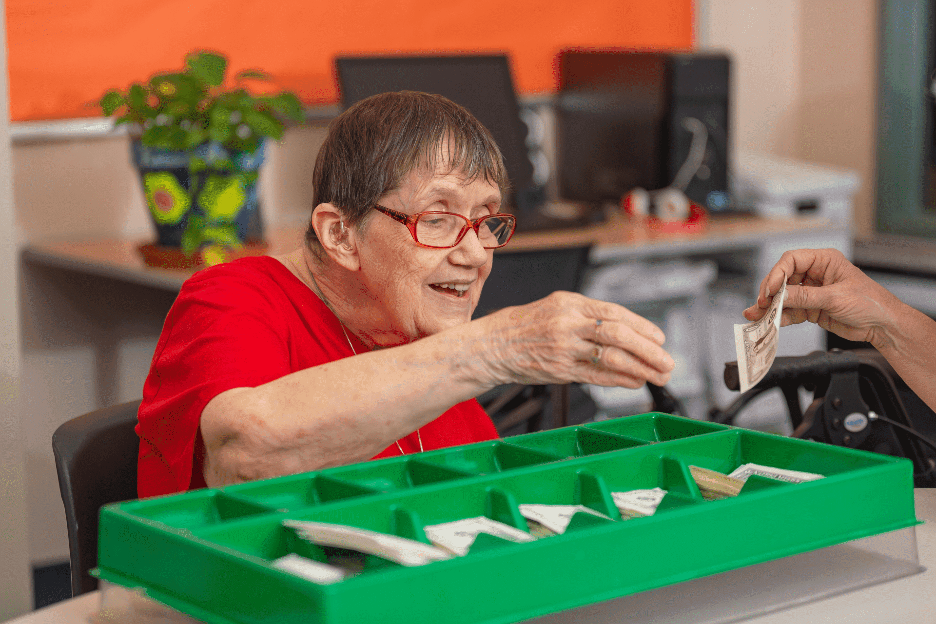 An elderly woman with glasses hands money to someone across a green tray with divided sections, in an early learning center setting with computers in the background.