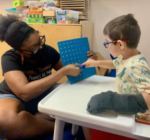 A woman in a mask provides pediatric therapy to a child at an early learning center. A woman in a mask provides pediatric therapy to a child at an early learning center.
