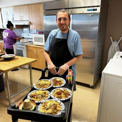 A man in an apron stands in a kitchen, smiling, next to a cart with several plates of food, as part of the adult day programs. Another person in a purple shirt is working diligently in the background. A man in an apron stands in a kitchen, smiling, next to a cart with several plates of food, as part of the adult day programs. Another person in a purple shirt is working diligently in the background.