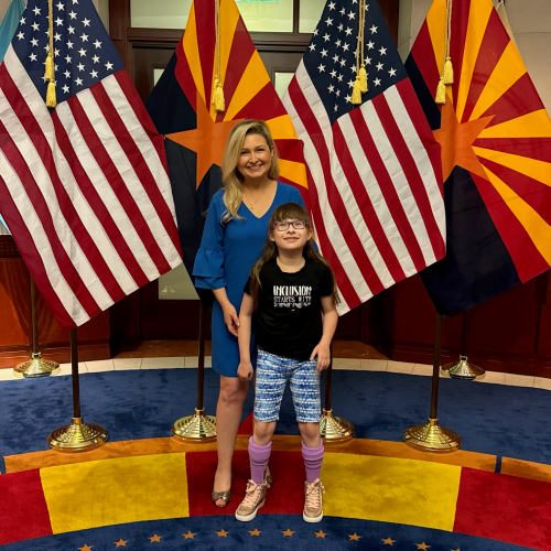 Two people stand smiling in front of three American flags and two Arizona state flags on flagpoles, indoors on a blue and red circular carpet.