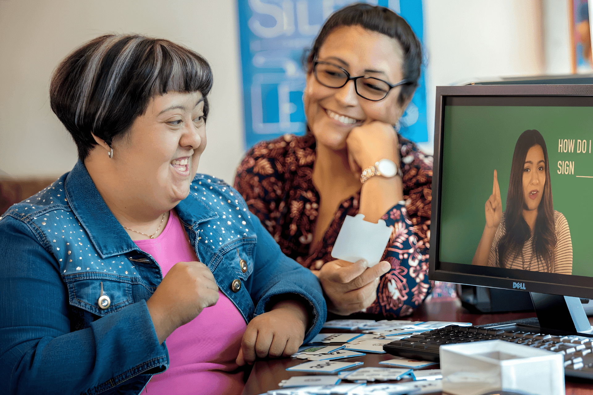 Two women sit together at a computer, smiling, while watching a sign language lesson video and holding flashcards.
