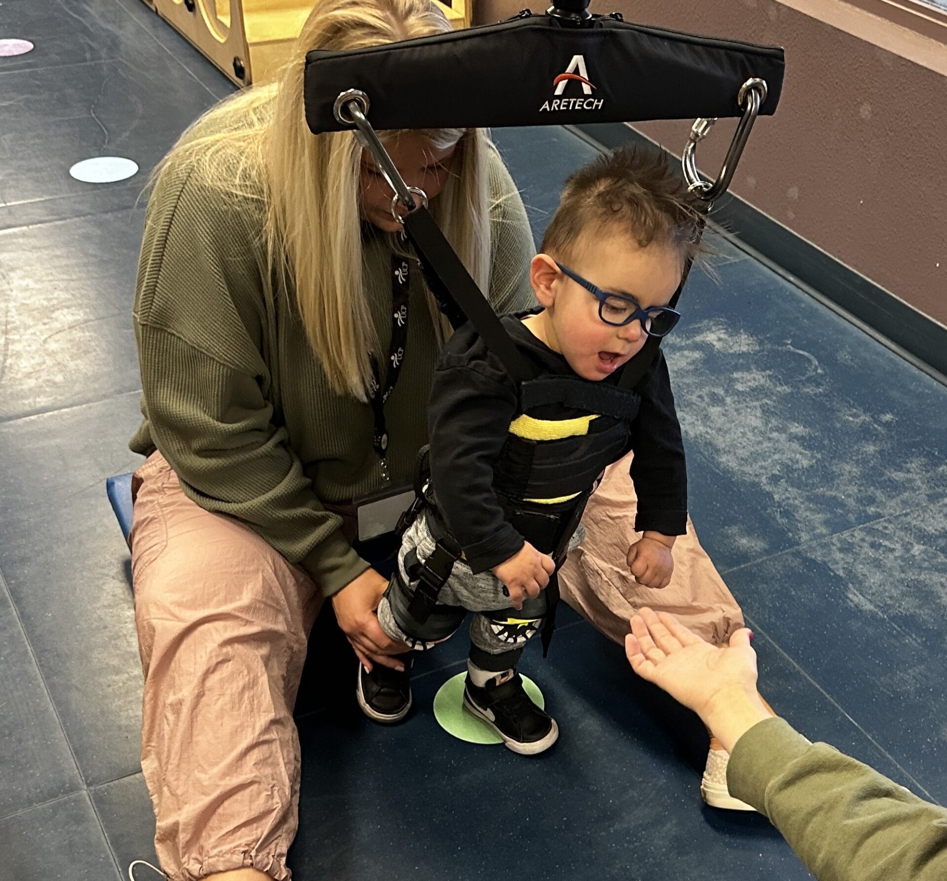 A child in a gait trainer is assisted by an adult during pediatric therapy while reaching out to a person's offered hand. Ace in a Zero G gait trainer in intensive therapy session