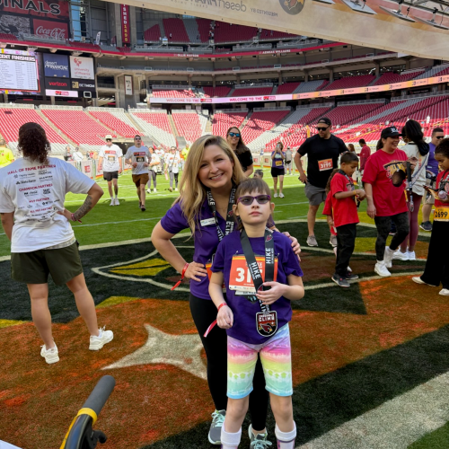 A woman and a girl with medals stand on a football field with other participants. The girl wears sunglasses and holds a medal; both are dressed in purple shirts.