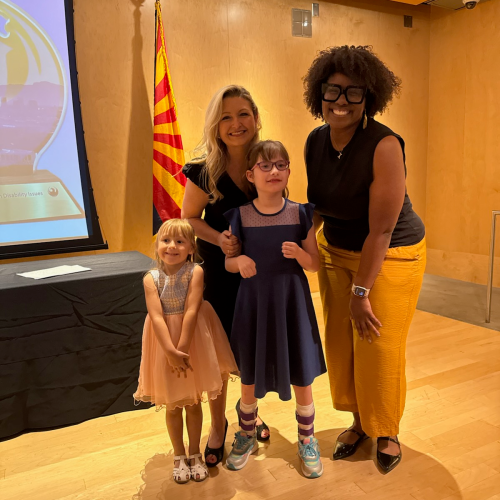 Two women and two young girls pose together indoors in front of a striped flag and a projector screen, smiling at the camera.
