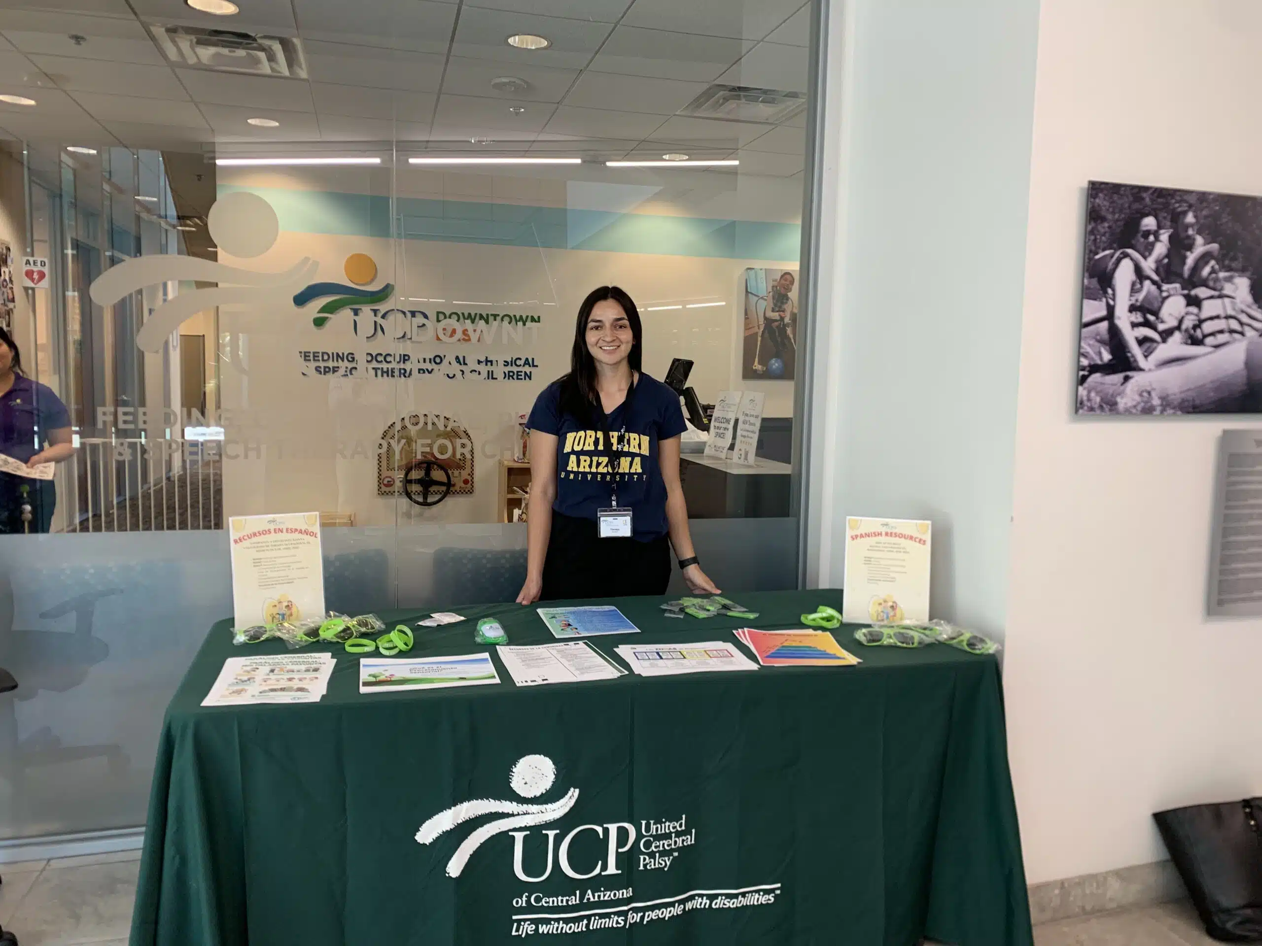 A woman stands behind a table with brochures and promo items, covered by a green UCP of Central Arizona tablecloth, in a building lobby with glass walls and informational displays, raising awareness about Cerebral Palsy. A woman stands behind a table with brochures and promo items, covered by a green UCP of Central Arizona tablecloth, in a building lobby with glass walls and informational displays, raising awareness about Cerebral Palsy.