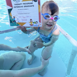 A young child wearing goggles smiles in a pool, holding a "Therapy Program Intensive Graduate" certificate. An adult supports the child from the side.