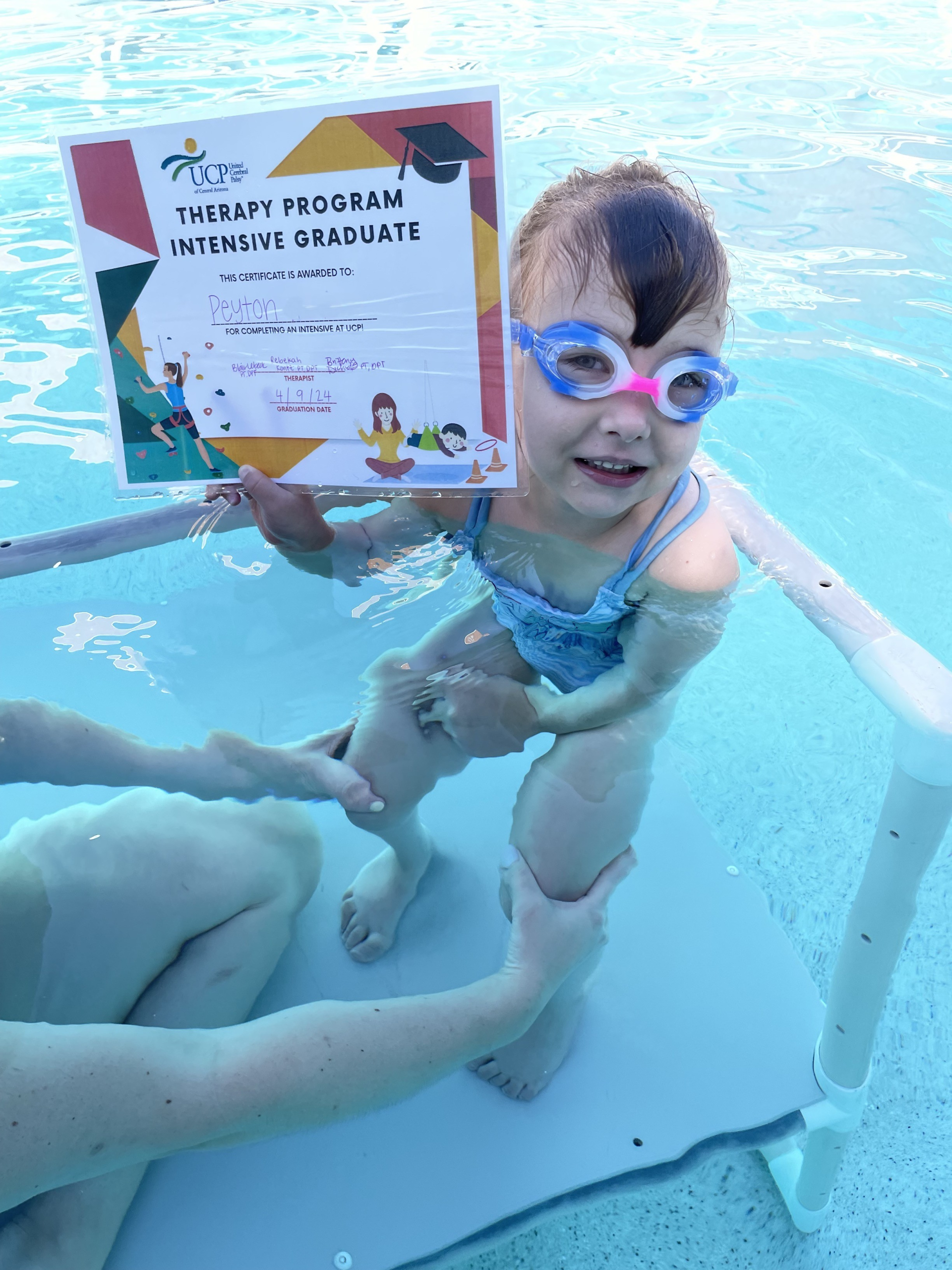 A young child wearing goggles smiles in a pool, holding a "Therapy Program Intensive Graduate" certificate. An adult supports the child from the side.
