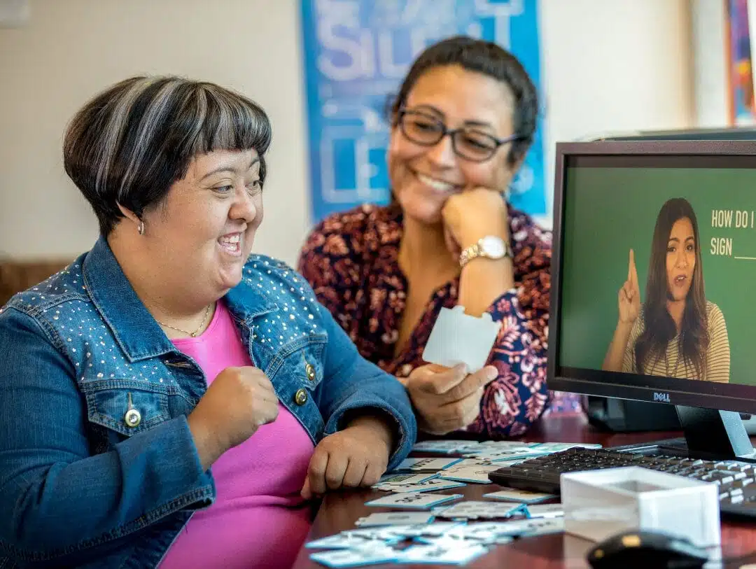 ArizonAbility: Adult Day Programs 2 Two women are seated at a desk, smiling and looking at a computer screen showing a woman using sign language. One woman, holding photo prints, is excitedly discussing the benefits of adult day programs for fostering inclusive communication.