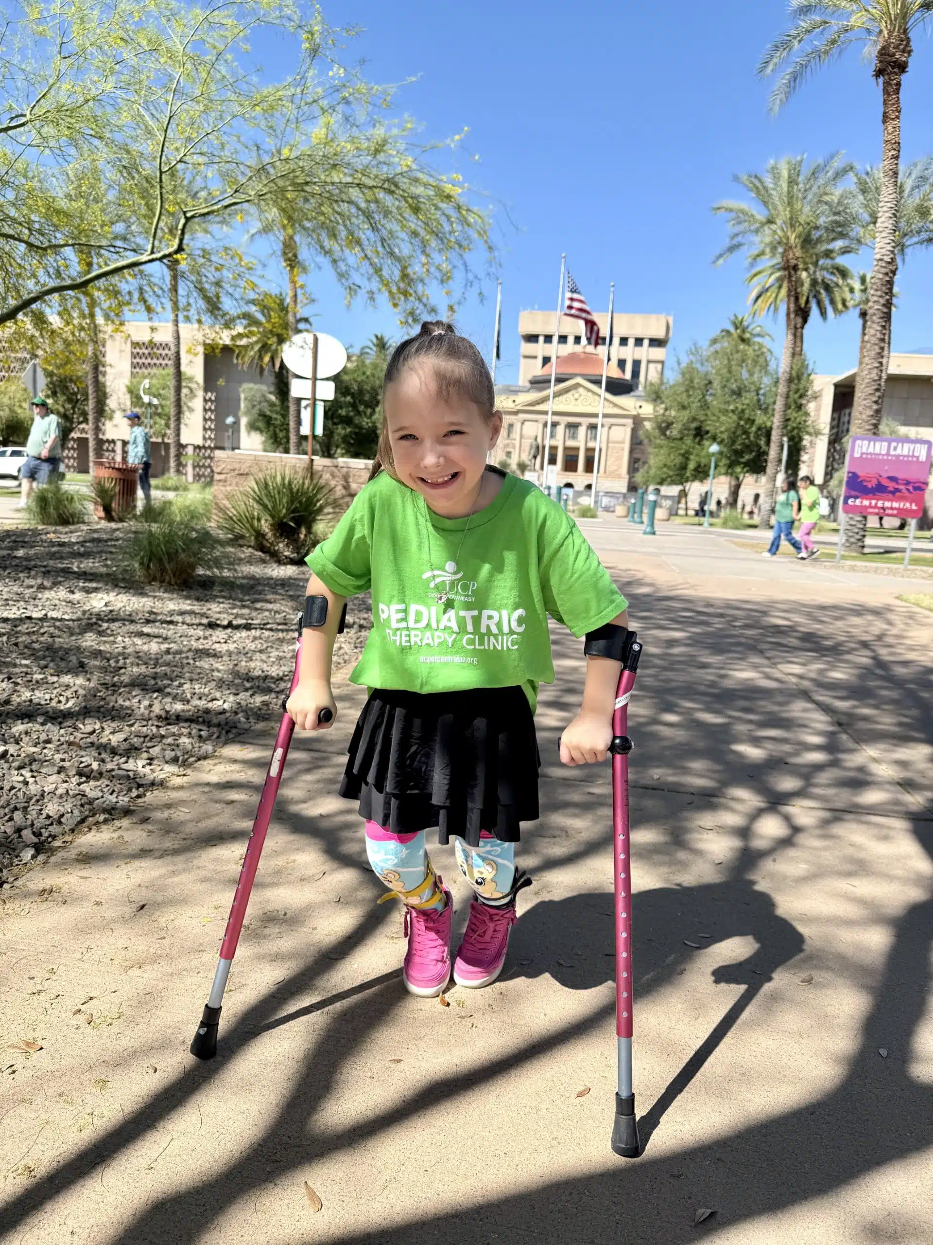Young girl with pink crutches smiles while standing on a sunny path, proudly wearing a green pediatric therapy clinic shirt that marks her as a UCP ambassador. She pairs it with a skirt and colorful leggings, while trees and buildings create a cheerful backdrop. Young girl with pink crutches smiles while standing on a sunny path, proudly wearing a green pediatric therapy clinic shirt that marks her as a UCP ambassador. She pairs it with a skirt and colorful leggings, while trees and buildings create a cheerful backdrop.