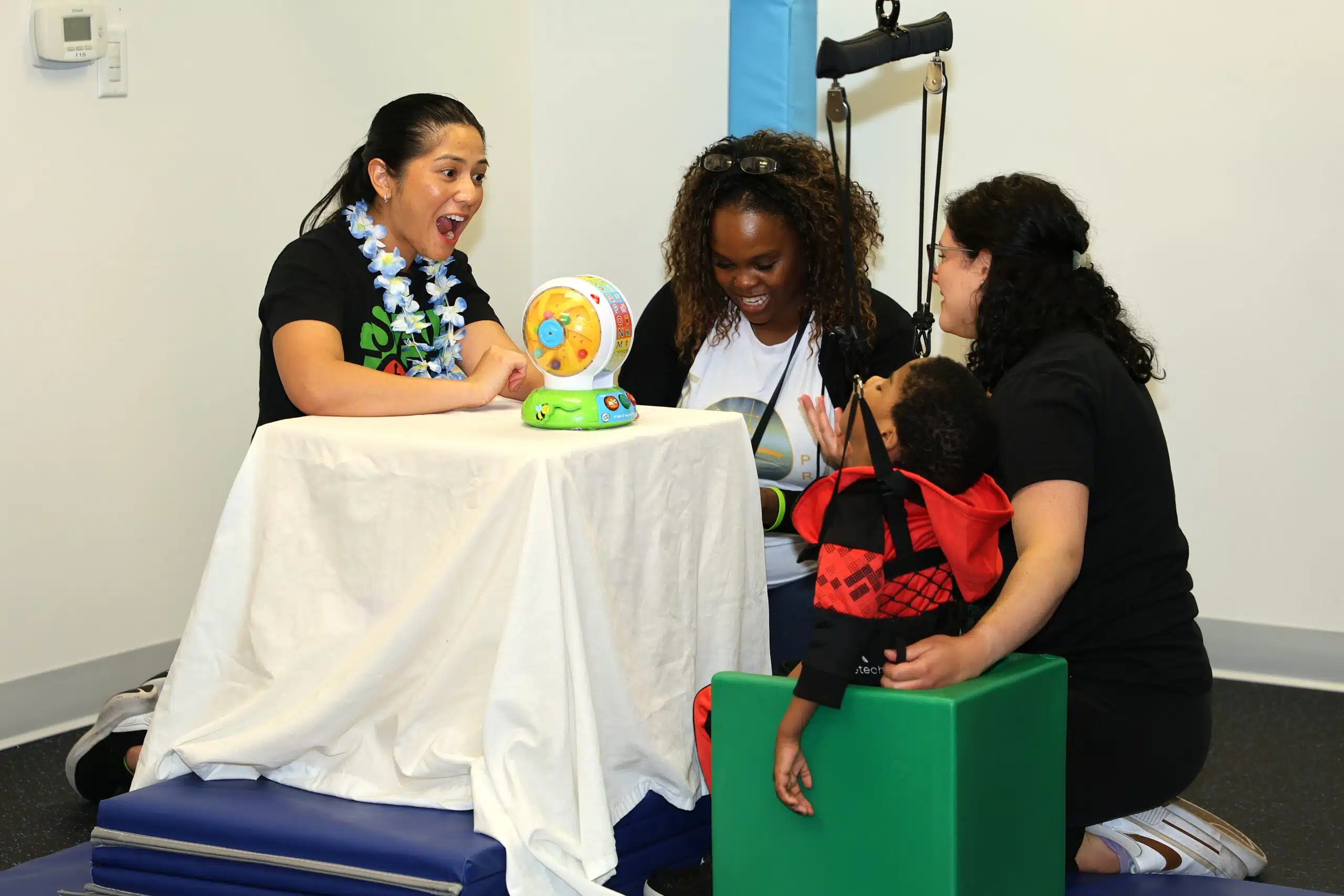Three women and a young boy interact around a small table with a toy in a brightly lit room, participating in a playful, therapeutic activity focused on early detection for Cerebral Palsy. Three women and a young boy interact around a small table with a toy in a brightly lit room, participating in a playful, therapeutic activity focused on early detection for Cerebral Palsy.
