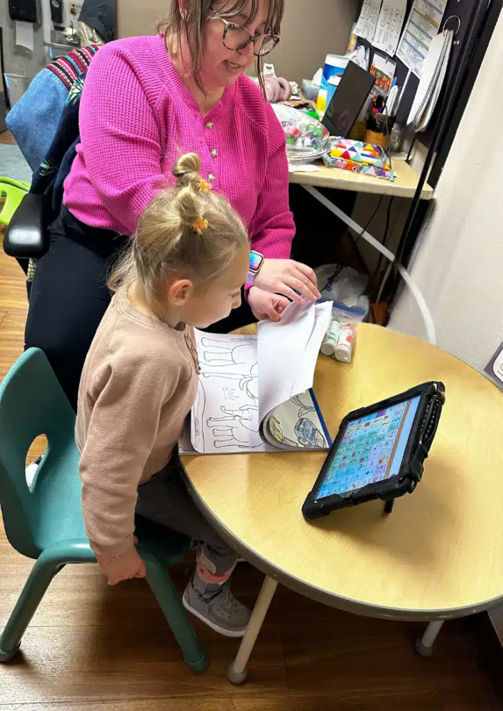 A young child sits at a table with a coloring book and an AAC communication device, while an adult in a pink sweater supervises beside her in a classroom or therapy setting. A young child sits at a table with a coloring book and an AAC communication device, while an adult in a pink sweater supervises beside her in a classroom or therapy setting.