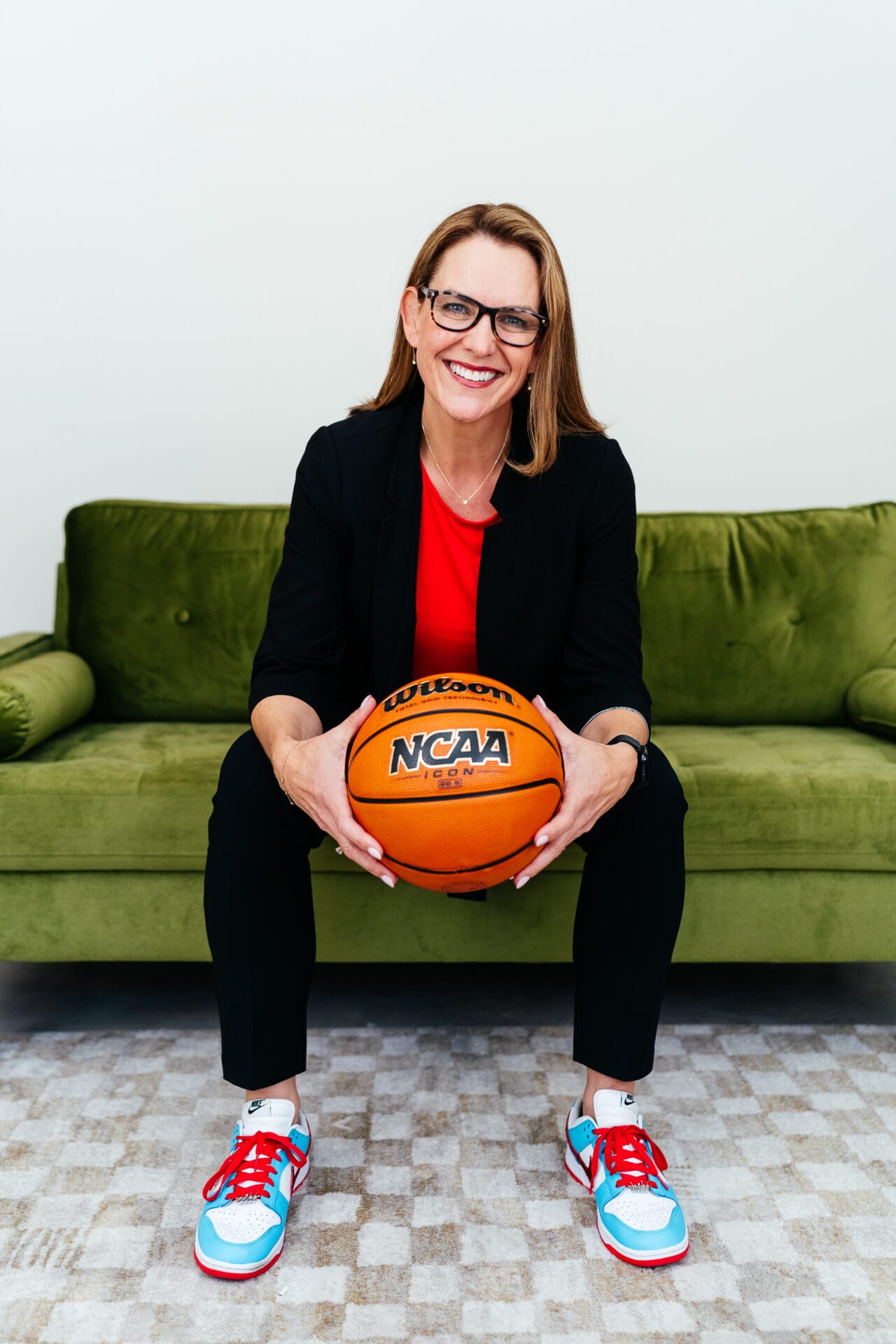 A person in glasses and a black suit sits on a green couch, holding an NCAA basketball, wearing red and blue sneakers. Sasha Graham UCP community ambassador. Podcast host of Sports Will Save Us All.