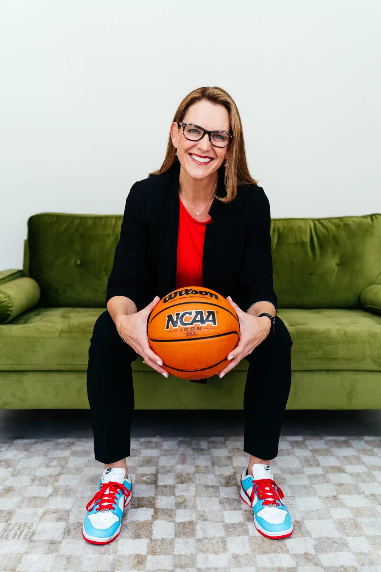 A person in glasses and a black suit sits on a green couch, holding an NCAA basketball, wearing red and blue sneakers. Sasha Graham UCP community ambassador. Podcast host of Sports Will Save Us All.