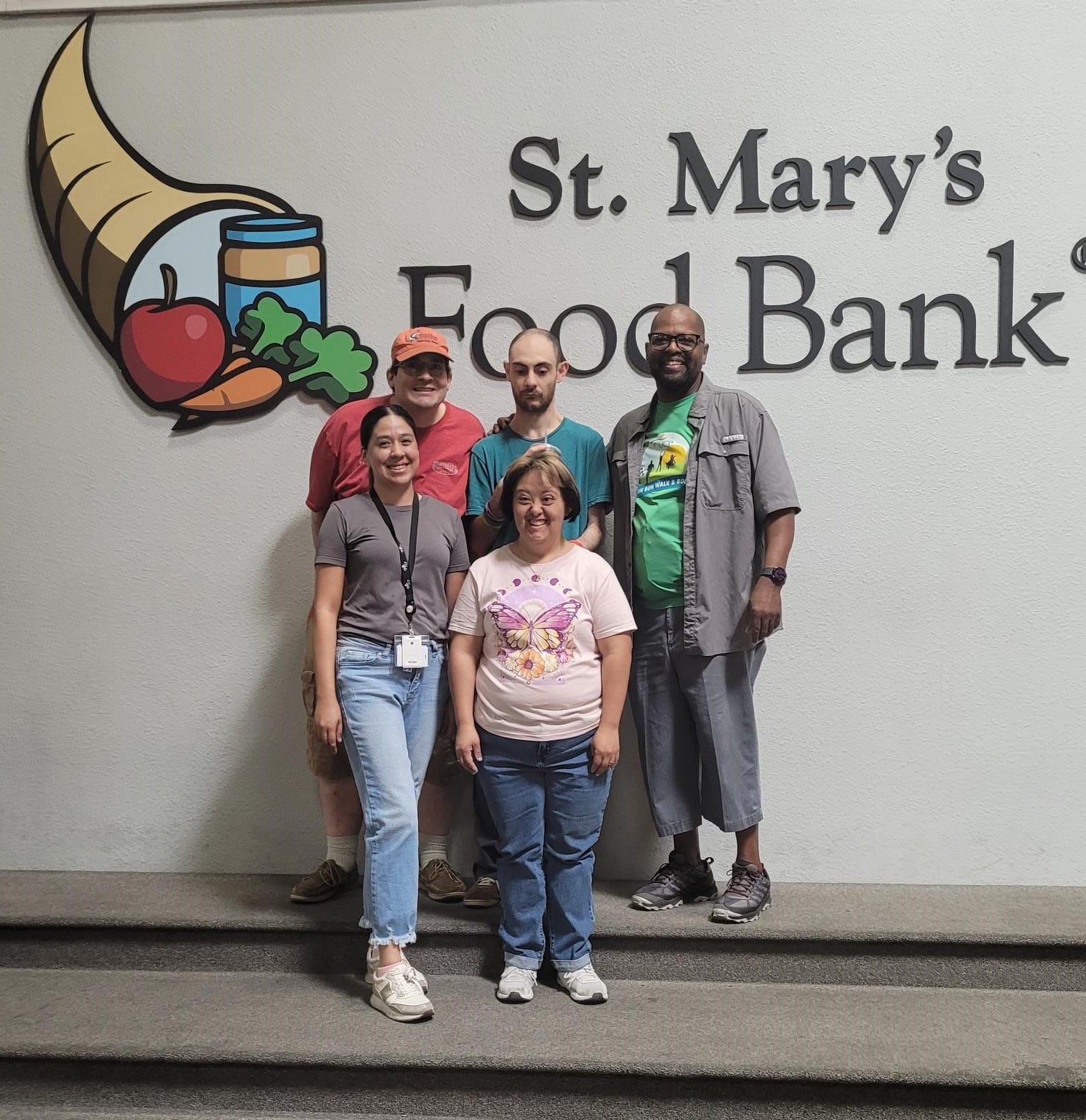 Five people stand in front of a wall with the logo and text "St. Mary's Food Bank." The group appears to be posing for a photo, with one member proudly displaying an AAC communication device. Five people stand in front of a wall with the logo and text "St. Mary's Food Bank." The group appears to be posing for a photo, with one member proudly displaying an AAC communication device.