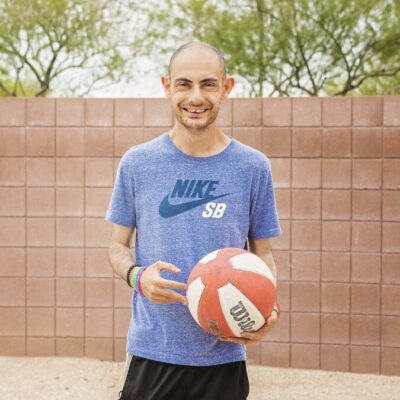 A person wearing a blue Nike SB shirt stands outside holding a red and white basketball, reminiscent of the recreational activities often found in adult day programs. There is a brick wall and trees in the background. A person wearing a blue Nike SB shirt stands outside holding a red and white basketball, reminiscent of the recreational activities often found in adult day programs. There is a brick wall and trees in the background.