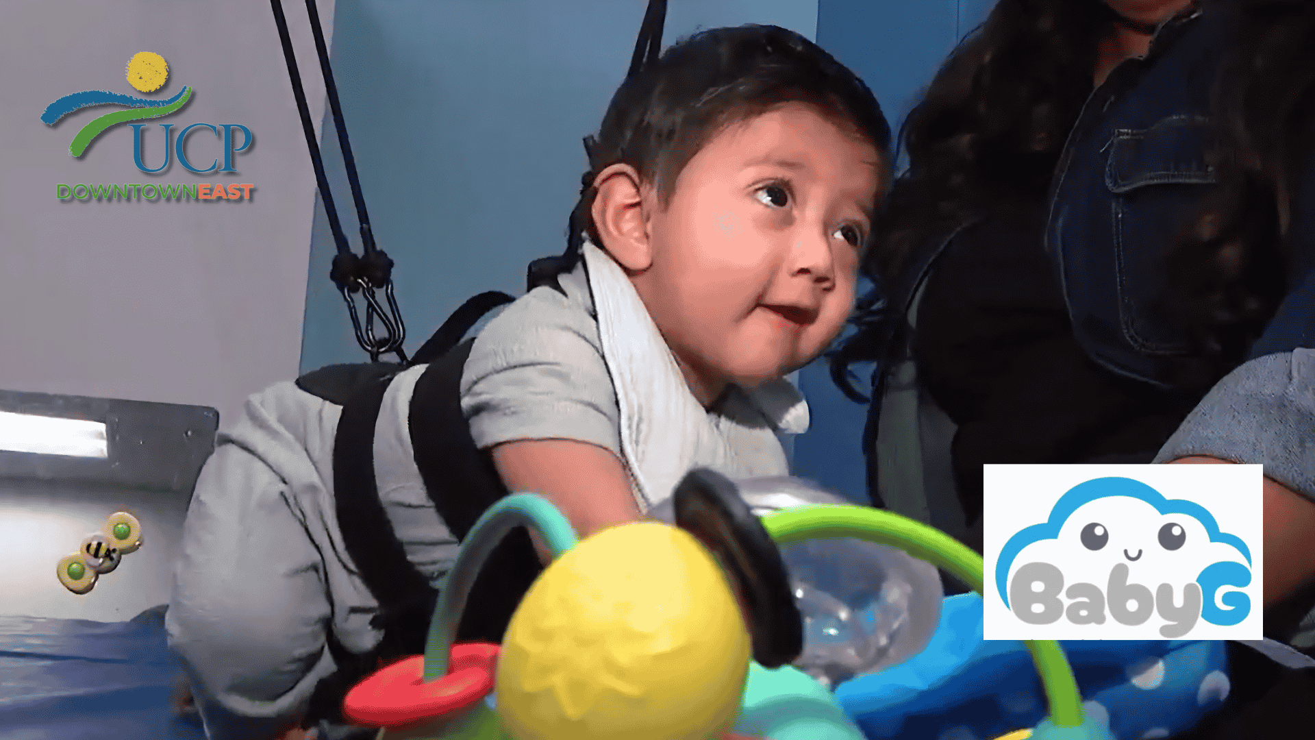 A baby in a harness is engaged in play therapy on a mat, with colorful toys in the foreground. UCP Downtown East and BabyG logos are visible on the image.
