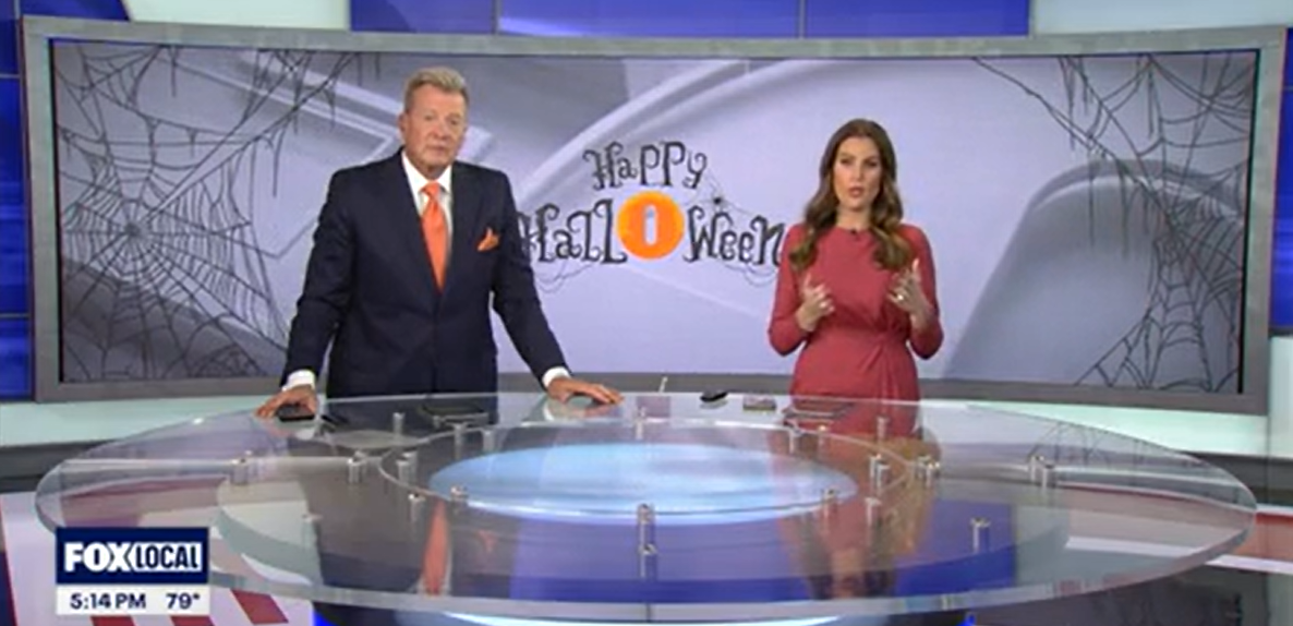 Two news anchors stand at a glass desk in a studio with a "Happy Halloween" graphic and spider webs displayed on the screen behind them, as they highlight UCP of central Az’s community events.