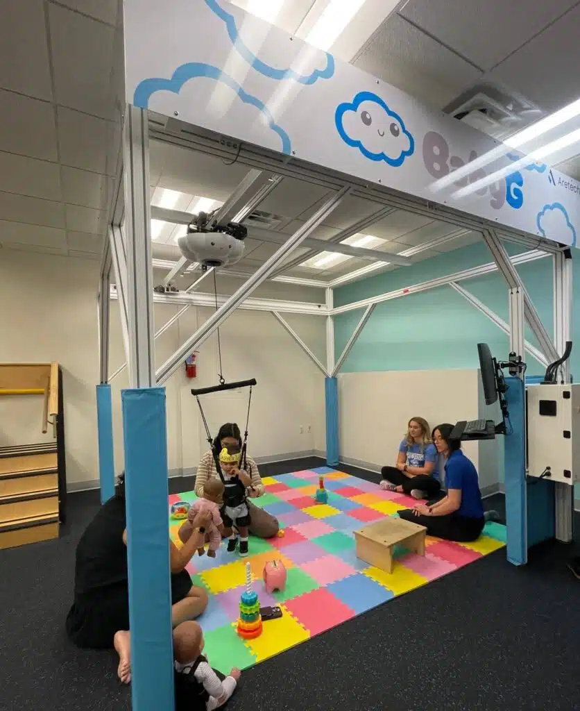 Four adults and one child interact on a colorful foam mat in a therapy room with play equipment and toys, supporting early detection for Cerebral Palsy, under a metal frame labeled "8iG. UCP of Central Az baby G robotic therapy session, supporting early detection for Cerebral Palsy, Baby G
