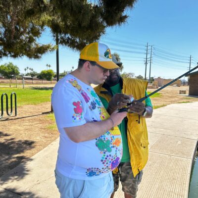 Two people stand outdoors, one in a colorful shirt and cap, the other in a yellow vest, working together to set up fishing equipment under a tree on a sunny day. It looks like an enjoyable activity from one of their adult day programs. Two people stand outdoors, one in a colorful shirt and cap, the other in a yellow vest, working together to set up fishing equipment under a tree on a sunny day. It looks like an enjoyable activity from one of their adult day programs.