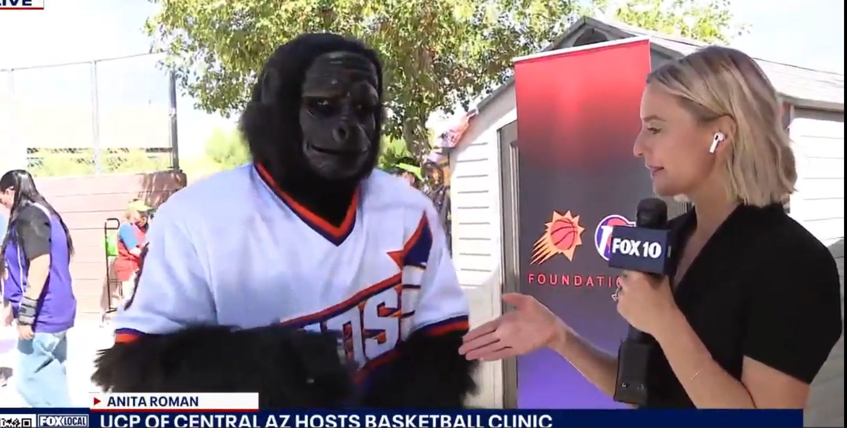 A woman holding a FOX 10 microphone interviews a person in a gorilla mascot costume wearing a basketball jersey at an outdoor event.