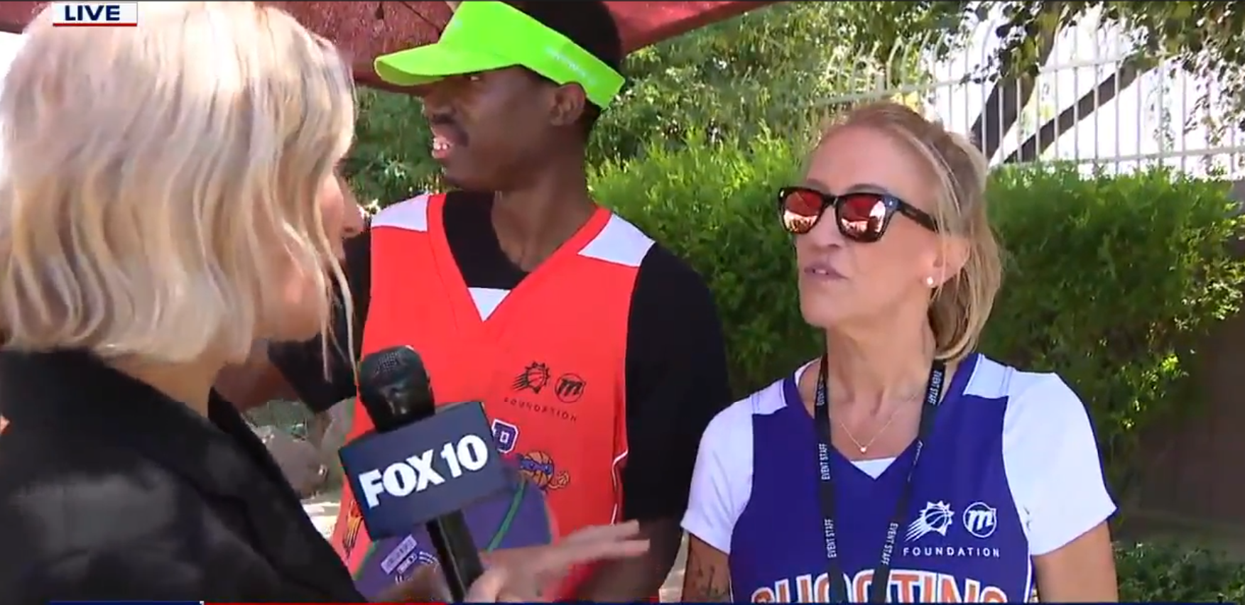 A reporter holding a FOX10 microphone interviews two people wearing sports jerseys outdoors.