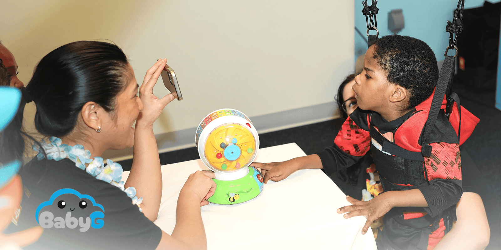 A child in a red and black harness interacts with a colorful spinning toy as an adult takes a photo with a smartphone. The BabyG logo is visible in the corner.