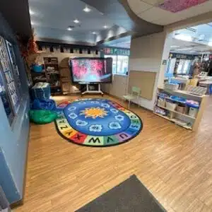 Early Learning Center 6 A cozy Early Learning Center classroom corner features a large screen, colorful alphabet rug, bean bags, small shelves with bins, and a green chair on a wood floor.