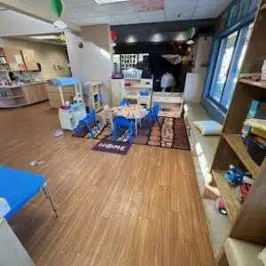 Early Learning Center 4 An Early Learning Center preschool classroom with small tables and chairs, play kitchen sets, shelves with toys, a rug, and a “HOME” mat on the floor.