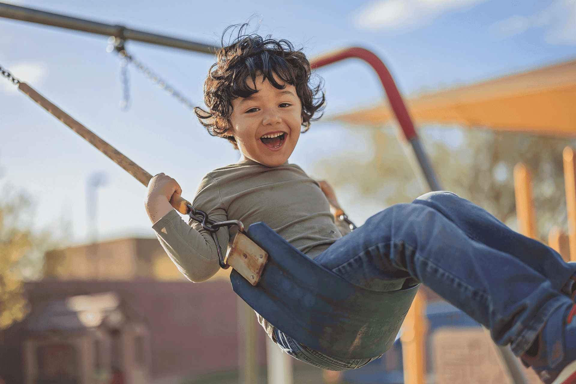 A young child with curly hair smiles while swinging at a playground on a sunny day.