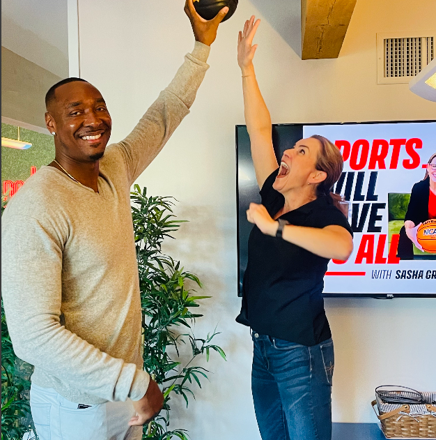 A man holds a kettlebell overhead while a woman, smiling and excited, reaches up toward it at UCP of Central AZ. In the background, a TV displays a sports show graphic.