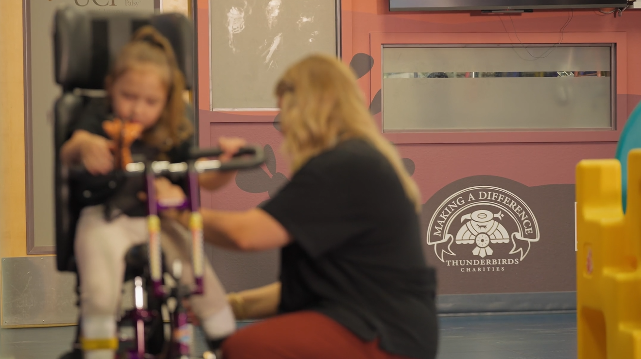 An adult assists a young child on a tricycle in an indoor setting at UCP of Central AZ, with a "Making a Difference" sign visible on the wall in the background.