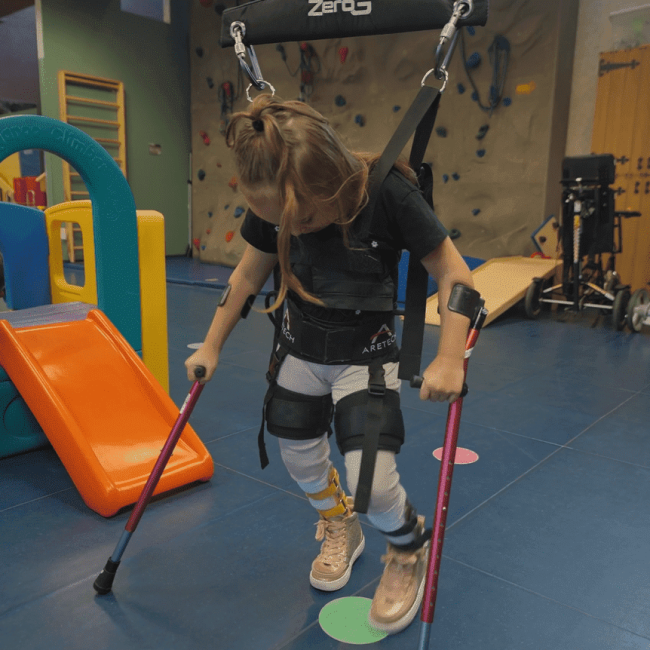 A young girl in a harness uses crutches for support in a therapy gym, with play equipment and a climbing wall in the background.