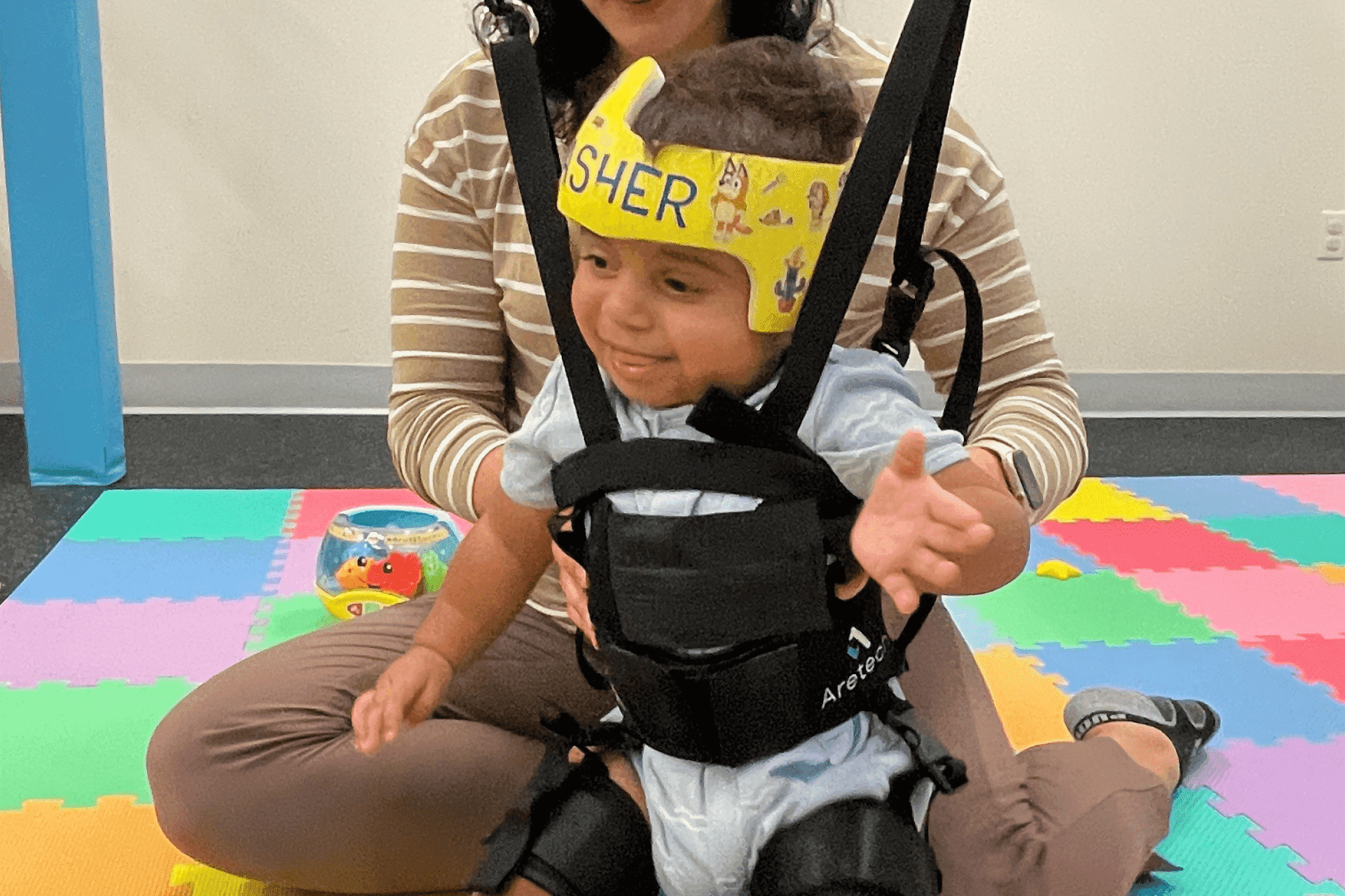A young child wearing a yellow headband and harness sits on a woman's lap on colorful foam mats in a playroom.
