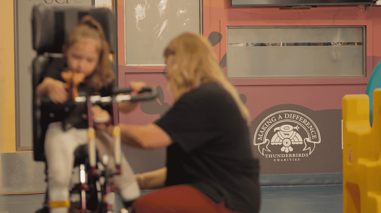A woman assists a young girl seated on a specialized tricycle in an indoor facility. A Thunderbirds Charities logo is visible on the wall in the background.