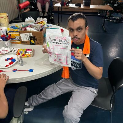 A man sits at a table holding up a piece of paper with colorful drawings labelled "13th of July Word Search". The table, part of the adult day programs, is cluttered with craft supplies, decorations, and other art materials. Arizonability visual arts table with colorful drawings labelled "4th of July Word Search".